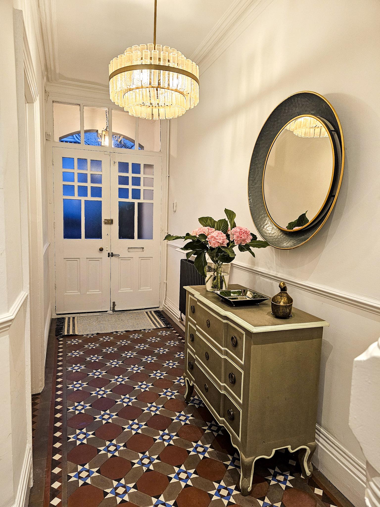 Hallway after restyling: the same tiled hallway lit warmly, with a painted chest of drawers, round mirror, pink hydrangeas and a runner by the door.